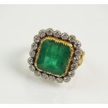 An emerald and diamond cluster ring, designed as a central trap cut emerald,