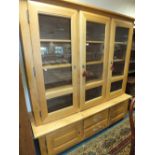 A light oak wall unit display cabinet with three glazed doors enclosing shelves above two short