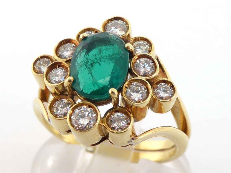 An emerald and diamond cluster ring, the central oval cut 8.4 x 6.6 x 3.2mm, in a surround of deep