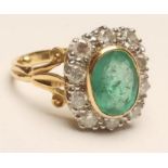 AN EMERALD AND DIAMOND CLUSTER RING, the oval facet cut emerald open back collet set within a border