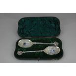 A cased pair of Victorian silver serving spoons with embossed scroll design, cherub mask finial