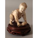 A JAPANESE IVORY OKIMONO OF A BOY, together with a fixed wood stand, the boy bending forward to lift