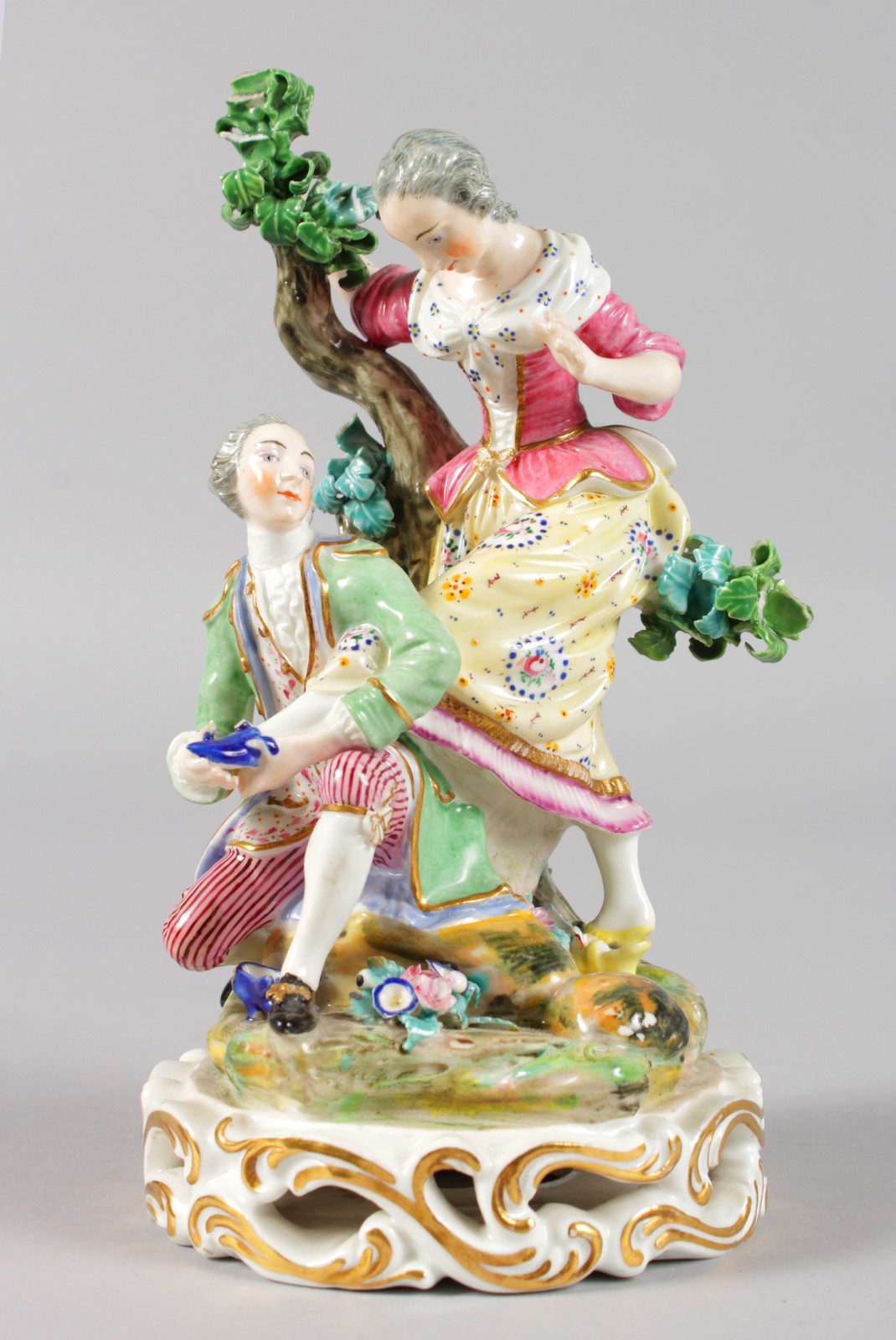 AN EARLY 19TH CENTURY DERBY FIGURE OF A SHOE BLACK, kneeling in front of a tree, holding a woman's