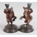 A PAIR OF PAINTED COLD CAST AMUSING FOX CANDLESTICKS on circular bases. 7ins high.