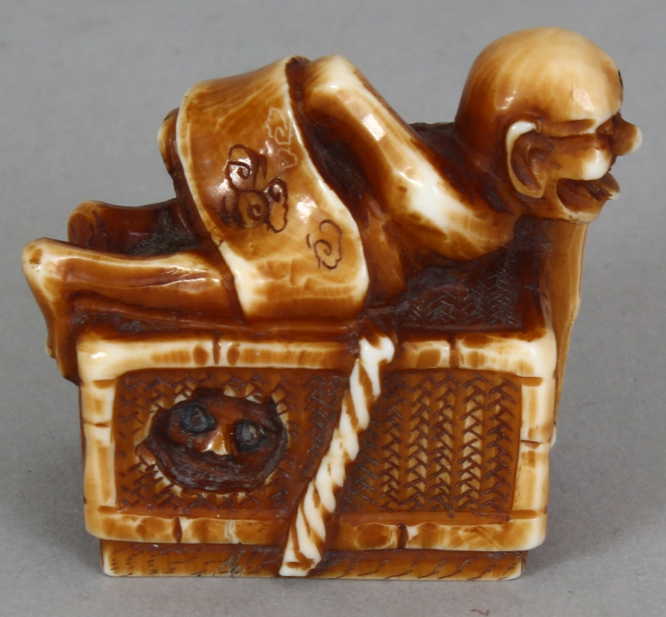 A SIGNED JAPANESE MEIJI PERIOD STAINED IVORY NETSUKE OF A DEMON, lying astride a rectangular reed