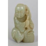 A CHINESE CELADON JADE FIGURE OF A BOY, seated next to a basket and supporting a fish on a leafy