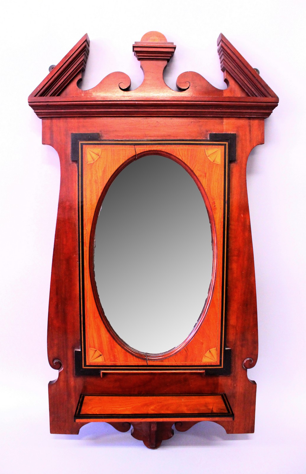 AN EDWARDIAN MAHOGANY FRAMED OVAL MIRROR with bevelled edge, broken arched pediment and small shelf.