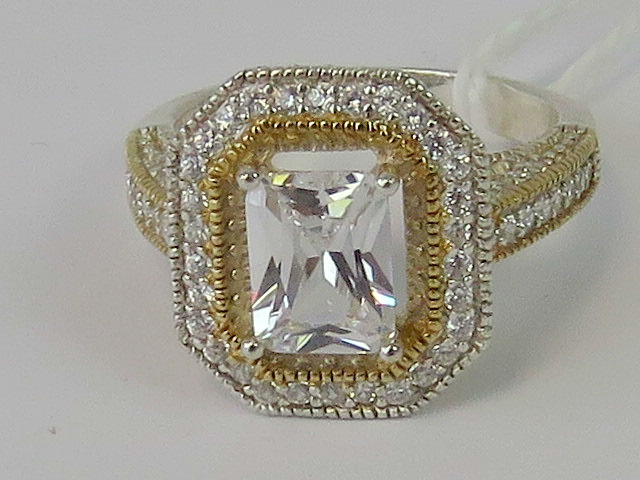 A silver halo style ring with central emerald cut white stone surrounded by a gallery of small