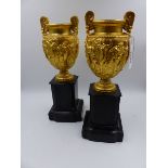 A PAIR OF 19th.C. GILT METAL CLASSICAL STYLE TWIN HANDLE URNS AFTER THE ANTIQUE WITH SHAPED BLACK