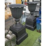 URNS, a pair, Victorian style in a black cast iron finish on plinths, 111cm x 60cm.