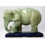 A GREEN GLAZED POTTERY ELEPHANT standing on a blue painted plinth base, 30cm x 26cm