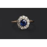 A SAPPHIRE AND DIAMOND CLUSTER RING, the circular mixed-cut sapphire claw set within a border of