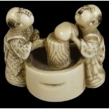 ˜A JAPANESE IVORY NETSUKE OF BOYS MAKING MOCHI, MEIJI PERIOD (1868-1912) standing either side of a