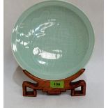 A Chinese celadon glazed plate decorated with lotus leaves, raised on a wood stand. 10'' diameter