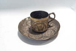 An Indian white metal cup and saucer, embossed with foliate scrolls and rural landscapes in panels,