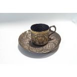 An Indian white metal cup and saucer, embossed with foliate scrolls and rural landscapes in panels,