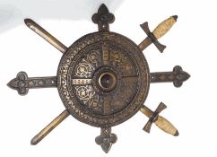 A cast iron wall plaque in the form of a shield in front of a Crucifix and two swords,