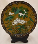 A Chinese cloisonné plate decorated with storks amongst foliage on an ochre ground raised on a