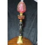 A VICTORIAN STYLE OIL LAMP the ruby glazed shade and reservoir supported by a black painted female