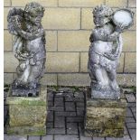 A pair of old reconstituted stone garden figures of putti playing a tambourine and cymbals on square