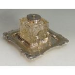 An Edwardian silver plated inkwell, the square glass well with sides cut with stylised suns,