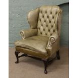 An olive green leather Chesterfield wing armchair raised on carved mahogany supports.