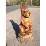 A chainsaw carved hare seated on his haunches, cut from an oak treetrunk with bark to plinth.