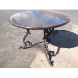 A circular garden table with hardwood top on three scrolled cast iron legs joined by circular