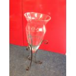 A tall tapering glass vase with flared rim, mounted in scrolled wrought iron tripod stand.