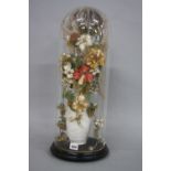 A VICTORIAN GLASS CYLINDRICAL DOME CONTAINING AN ARRANGEMENT OF WOVEN SILK FLOWERS AND REAL GRASSES,
