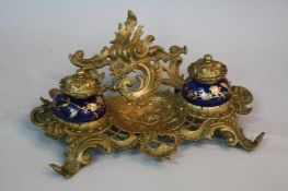 A LATE VICTORIAN BRASS DESK STAND, cast with foliate scrolls, the two ceramic inkwells marked for