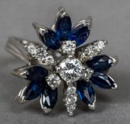 An unmarked white gold diamond and sapphire ring Of floral cluster form. 2 cm wide.