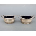 A pair of hallmarked silver two-handled open salts with blue glass liners, Sheffield 1907 and 1910,