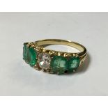 A YELLOW METAL, EMERALD AND DIAMOND RING With a single oval cut diamond flanked by four emeralds and