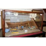 Cased diorama of a boatyard with wooden hull under construction