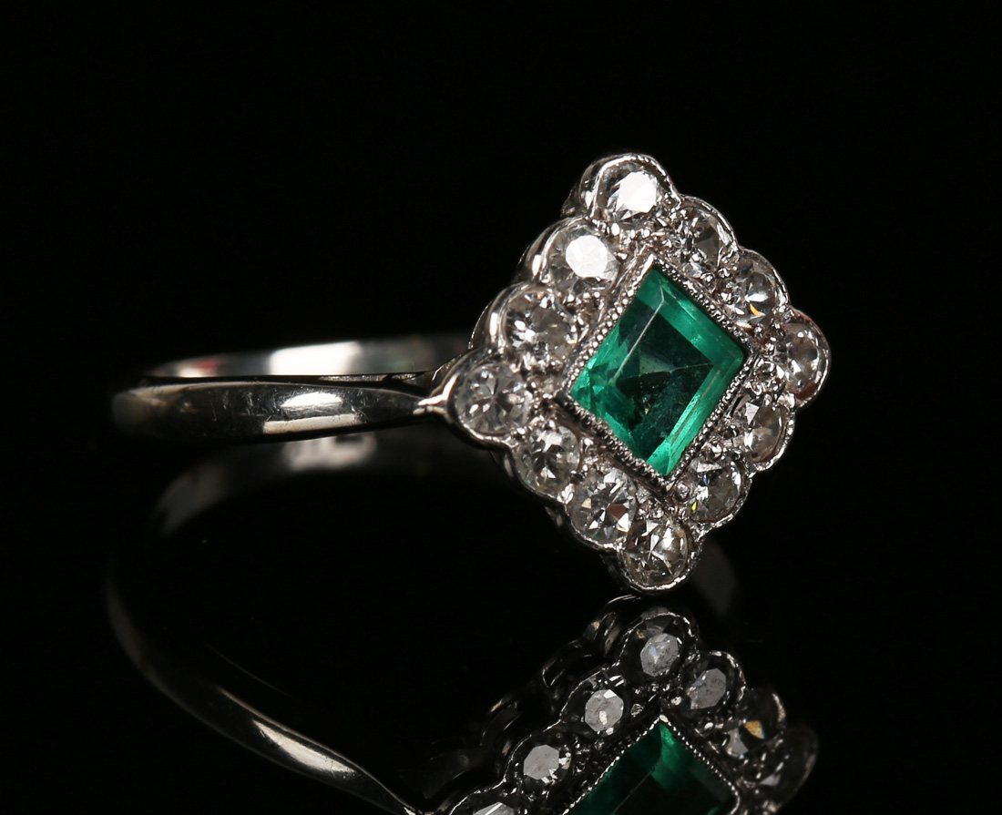 A platinum, emerald and diamond lozenge shaped cluster ring, mounted with the square cut emerald