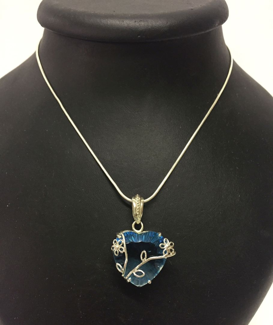 A large pendant on a silver chain with a heart shaped blue stone and flower decoration. Bale