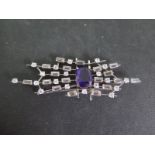 A white metal brooch set with a central Amethyst surrounded by white stones in a modernist setting