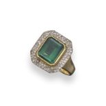 An emerald and diamond cluster ring, the emerald-cut emerald is set within a surround of small