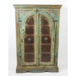 A painted hard wood two door cabinet, with carved arched doors enclosing shelves, on plinth base,