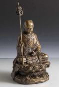 Figurine, Buddha, Bronze, auf Lotussockel sitzend in ornamental verziertem Umhang, einen Stab