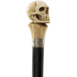 AN EARLY 20TH CENTURY WALKING CANE, the ivory handle carved as a skull, above silver collar