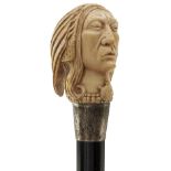 AN EARLY 20TH CENTURY WALKING CANE, the ivory pommel carved as the head of a Native American in