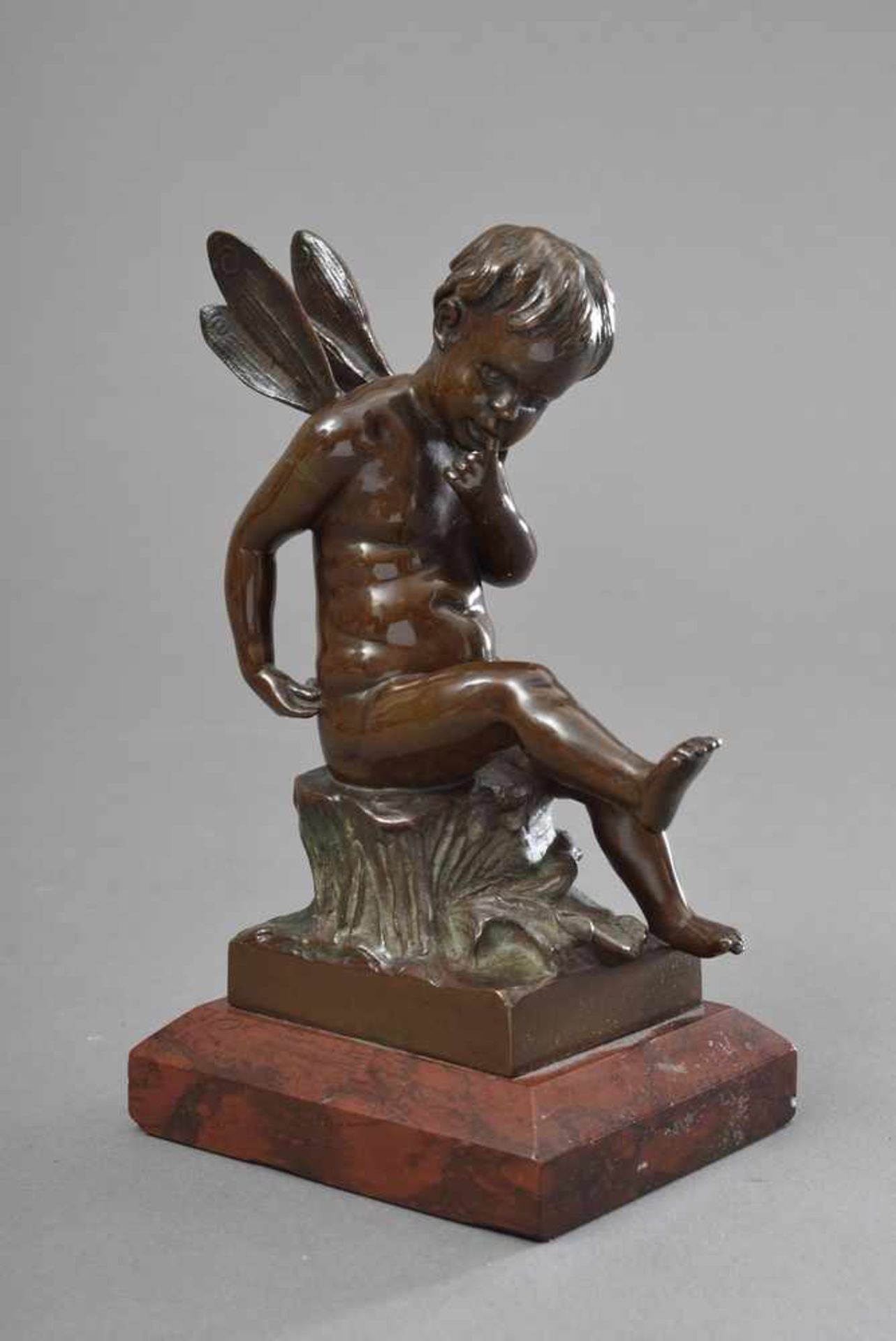 Kleine Bronze "Sitzende Amourette mit Insektenflügeln" auf rotem Steinsockel, Gießereisignet "H.