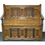 An Old Charm oak hall settle, carved panel back, open arms, hinged seat, carved panel front.