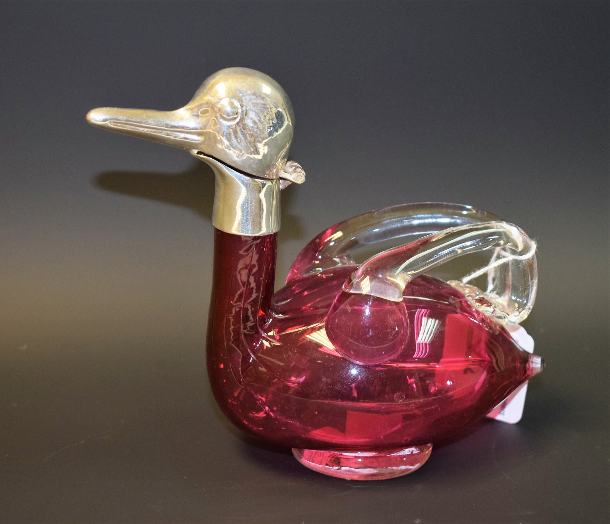 A cranberry glass duck shaped claret jug, white metal mounts,