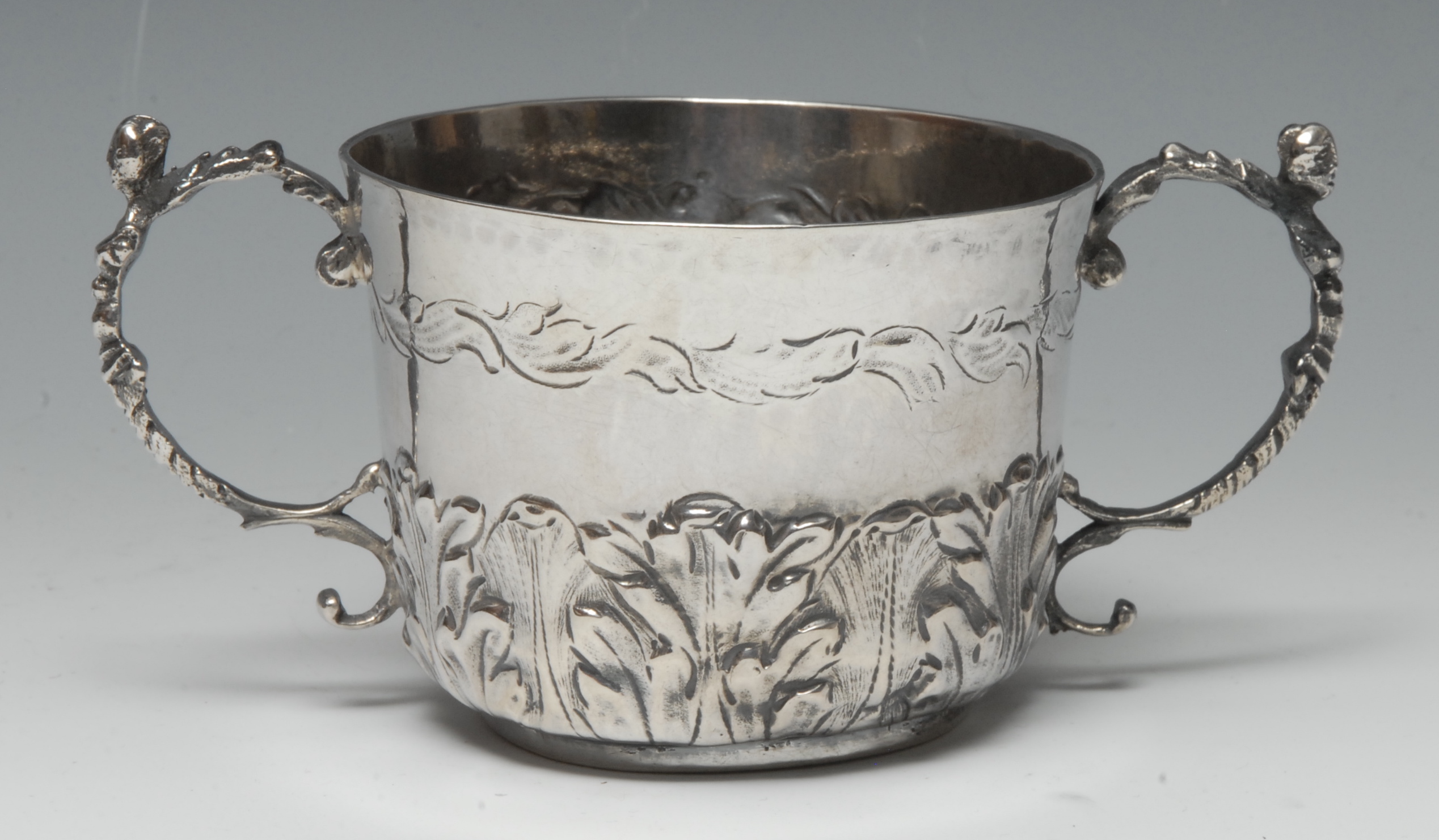 A Charles II silver porringer, chased with bands of acanthus, scroll handles capped with masks,