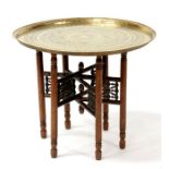 An Islamic brass tray-top table, on folding hardwood base, 69cms (27.25ins) diameter.