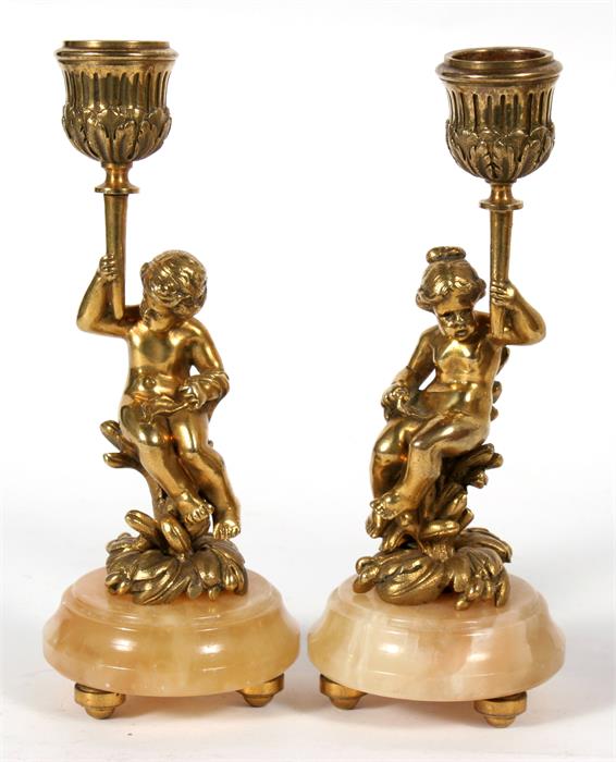 A pair of bronze figural candlesticks depicting young children sat on a flowering plant, 17cms (6.