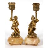 A pair of bronze figural candlesticks depicting young children sat on a flowering plant, 17cms (6.